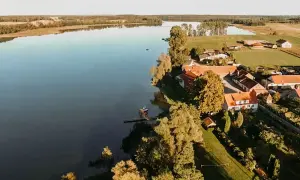 Malownicze jezioro Dejguny z zabudowaniami i zielonymi polami. Widać małą łódkę na wodzie i pomost.