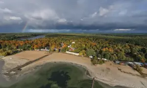 Jesioro Niedzięgiel w jesiennej odsłonie. Tęczowy łuk nad ciemnymi chmurami, piaszczysta plaża i kolorowe drzewa tworzą malowniczy krajobraz.