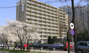 Hotel Felix Warszawa w otoczeniu kwitnących drzew. Widać parking z samochodami i budynek hotelu z balkonami.