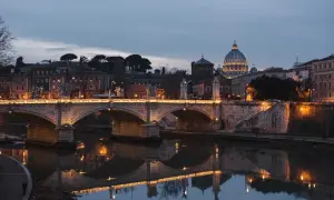 Most Ponte Sant'Angelo, oświetlony nocą, odbija się w spokojnej wodzie rzeki Tyber. W tle majaczy kopuła Bazyliki św. Piotra.