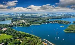 Malownicze mazury miejscowości nad jeziorem, z zielonymi lasami, białymi żaglami i błękitnym niebem.