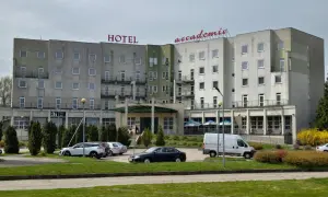 Hotel Accademia w Przemyślu, budynek z czerwonym napisem, parking z samochodami i zielony trawnik.