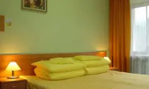 Pokój w sanatorium Mielec Krynica Zdrój. Jasne ściany, łóżko z żółtą pościelą i poduszkami, lampka nocna, pomarańczowa zasłona.