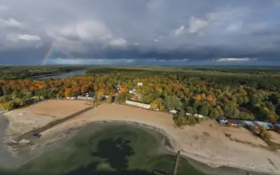 Jesioro Niedzięgiel w jesiennej odsłonie. Tęczowy łuk nad ciemnymi chmurami, piaszczysta plaża i kolorowe drzewa tworzą malowniczy krajobraz.