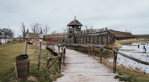 Drewniany mostek prowadzi do grodziska z wieżą strażniczą i zabudowaniami, część szlaku piastowskiego.