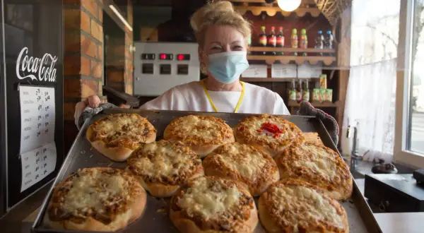 Kobieta w maseczce prezentuje blachę z pizzami. To może być najstarsza pizzeria w Polsce, gdzie tradycja pieczenia pizzy trwa od lat.