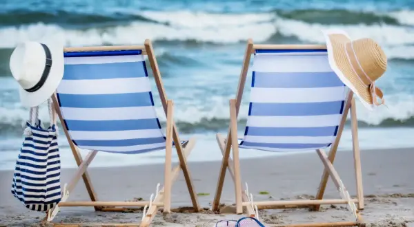 Dwa leżaki plażowe z niebiesko-białymi pasami, kapelusze i torba na piasku, z widokiem na morze.