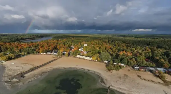 Jesioro Niedzięgiel w jesiennej odsłonie. Tęczowy łuk nad ciemnymi chmurami, piaszczysta plaża i kolorowe drzewa tworzą malowniczy krajobraz.