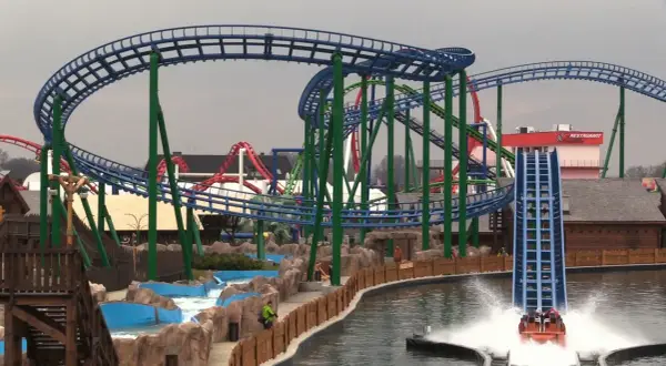 Water coaster zjeżdża z dużej wysokości, tworząc fontannę wody. W tle widać inne atrakcje parku rozrywki.