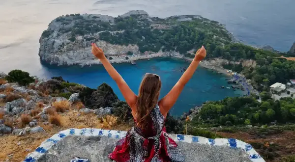 Kobieta z rękami w górze podziwia zatokę. Nawet w listopadzie na Rodos pogoda sprzyja pięknym widokom.
