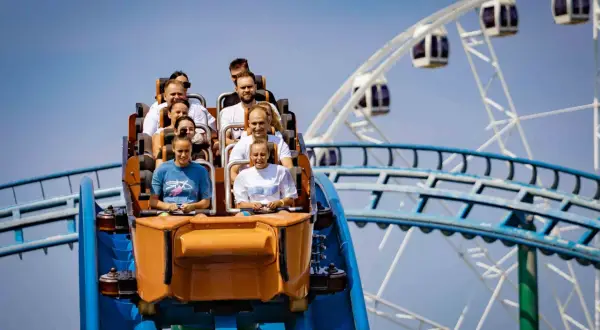 Ludzie na wagoniku rollercoastera przeżywają emocje podczas zjazdu. W tle widać koło widokowe.