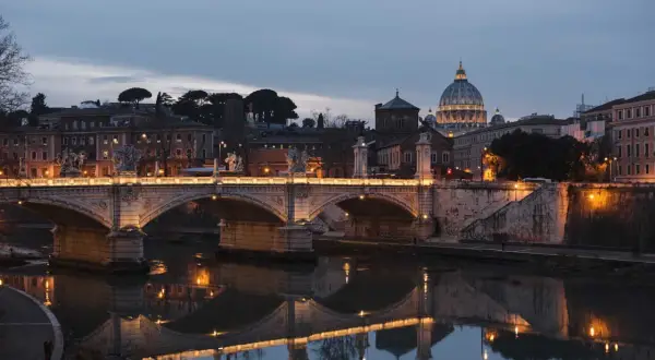 Most Ponte Sant'Angelo, oświetlony nocą, odbija się w spokojnej wodzie rzeki Tyber. W tle majaczy kopuła Bazyliki św. Piotra.
