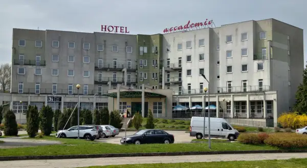 Hotel Accademia w Przemyślu, budynek z czerwonym napisem, parking z samochodami i zielony trawnik.