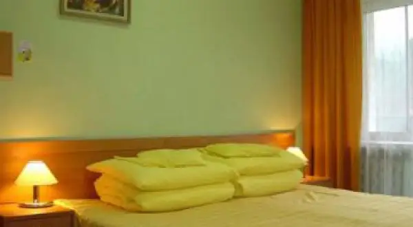 Pokój w sanatorium Mielec Krynica Zdrój. Jasne ściany, łóżko z żółtą pościelą i poduszkami, lampka nocna, pomarańczowa zasłona.