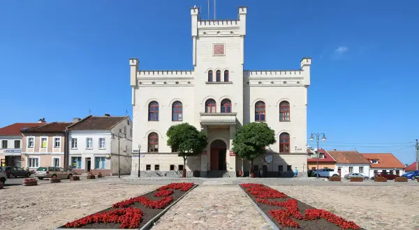 Jasny ratusz z wieżą i czerwonymi rabatami kwiatów na brukowanym placu. Pasym atrakcje turystyczne zachęcają do zwiedzania.