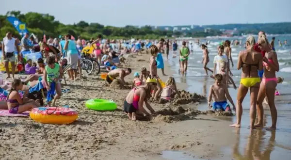 Tłum ludzi cieszy się słońcem i zabawą na plaży w Gdańsku. Dzieci budują zamki z piasku, a dorośli odpoczywają nad morzem.