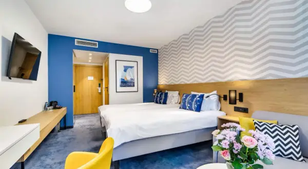 Pokój w hotelu Mistral Jurata: dwa łóżka, żółty fotel, kanapa, bukiet kwiatów i obraz z żaglówką.