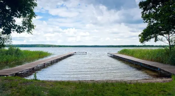 Dwa drewniane pomosty prowadzące na spokojne jezioro, otoczone zielenią. Idealne miejsce na relaks i podziwianie przyrody, jedna z wielu atrakcji w Boszkowie.