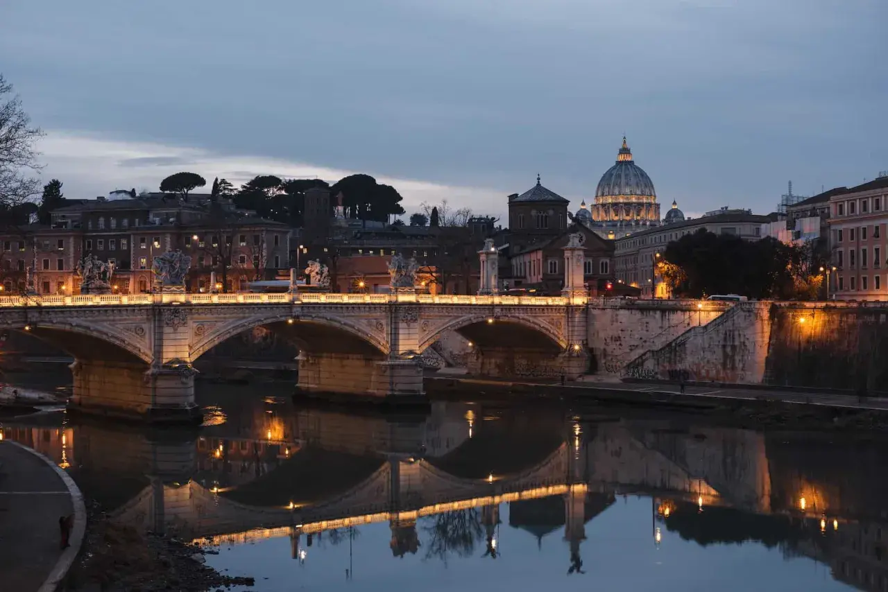 Most Ponte Sant'Angelo, oświetlony nocą, odbija się w spokojnej wodzie rzeki Tyber. W tle majaczy kopuła Bazyliki św. Piotra.