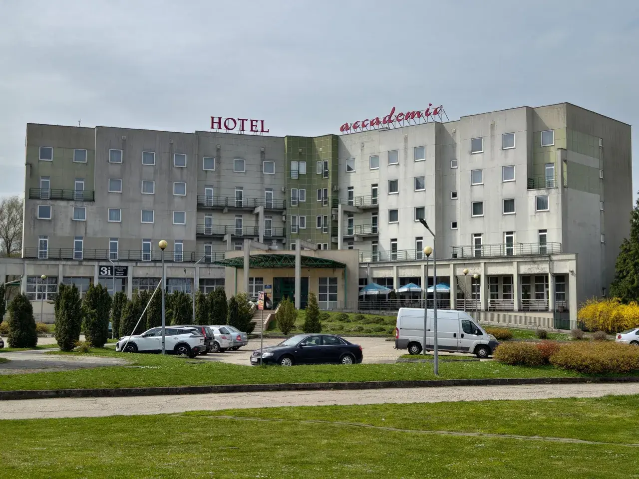 Hotel Accademia w Przemyślu, budynek z czerwonym napisem, parking z samochodami i zielony trawnik.