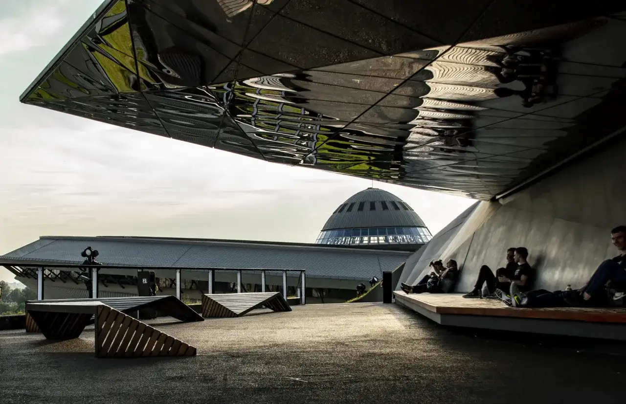 Nowoczesna architektura i nietypowe atrakcje Katowice. Ludzie odpoczywają pod futurystycznym zadaszeniem, w tle kopuła Muzeum Śląskiego.