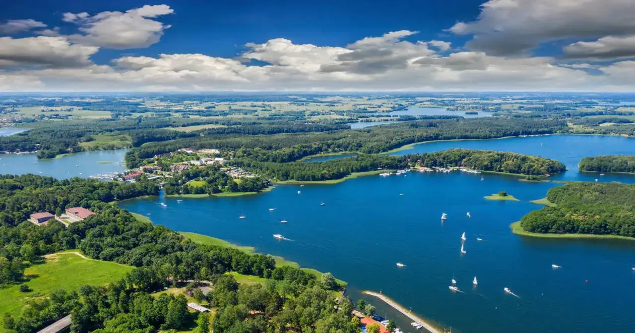 Malownicze mazury miejscowości nad jeziorem, z zielonymi lasami, białymi żaglami i błękitnym niebem.