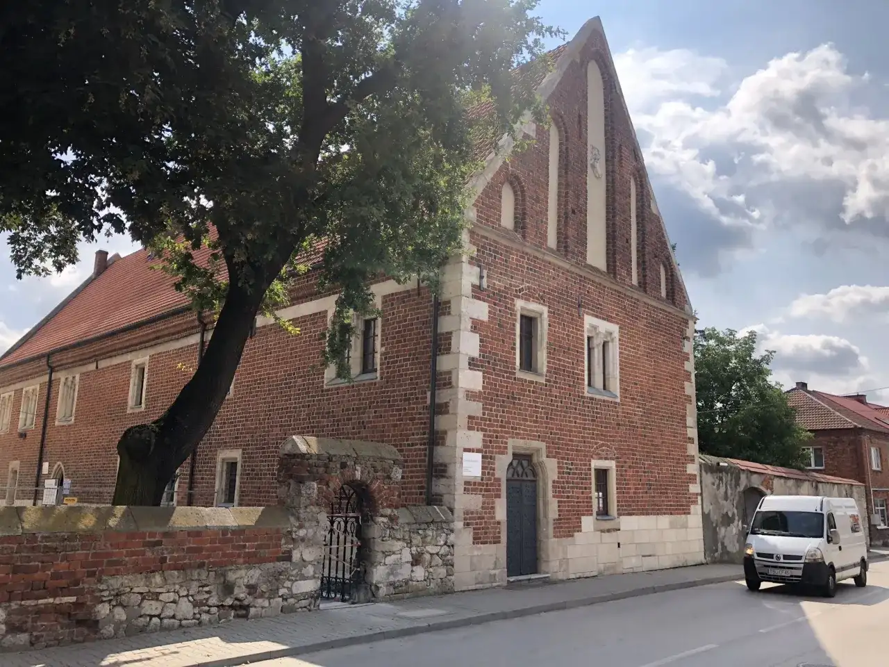Kamienica z cegły, ozdobiona kamiennymi detalami, to jedna z kazimierza wielka atrakcje. Obok rośnie stare drzewo.