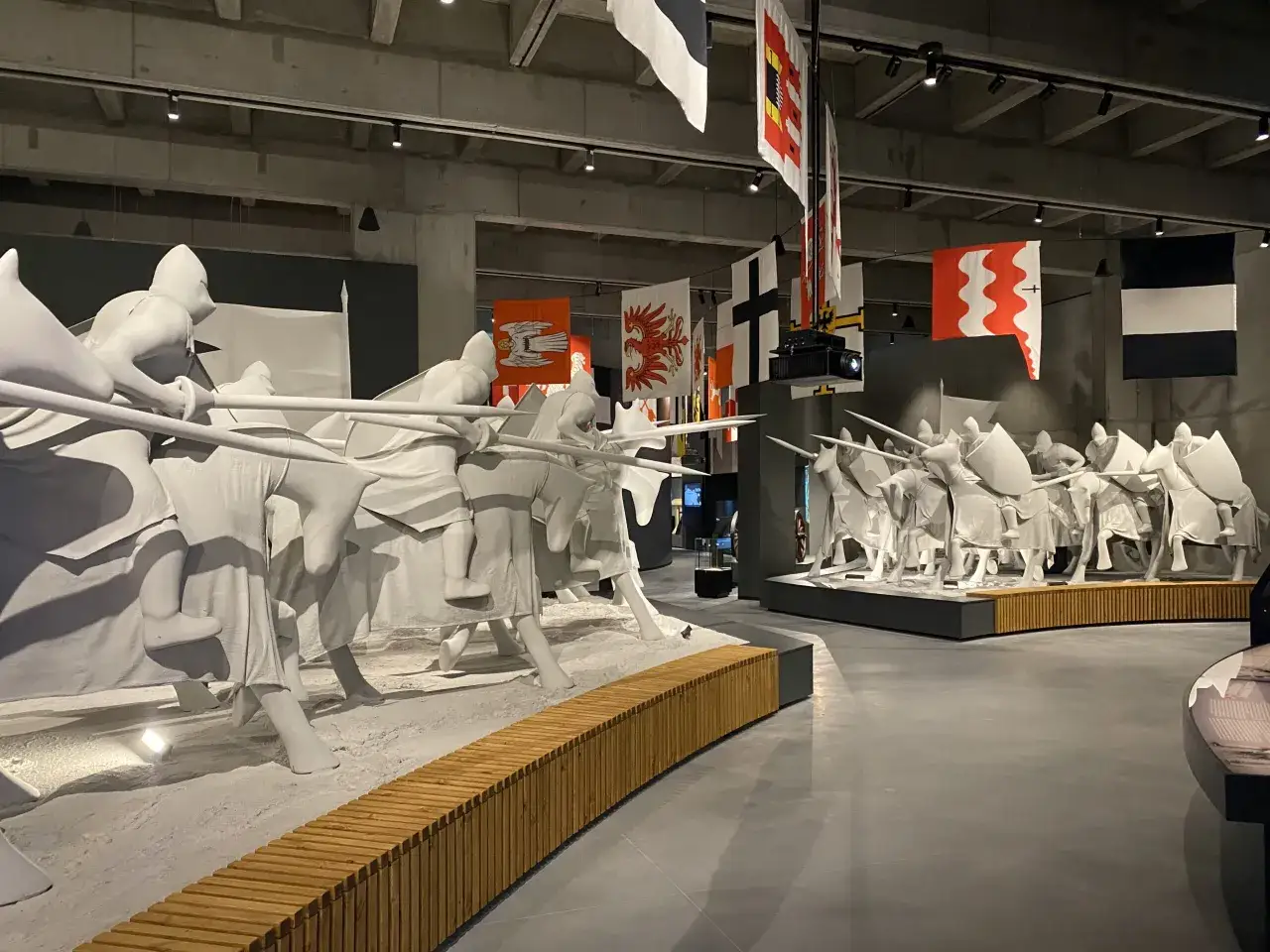 Białe rzeźby rycerzy na koniach, nawiązujące do bitwy pod Grunwaldem. W tle wiszą historyczne flagi, tworząc atmosferę ekspozycji. To jedne z wielu atrakcji w muzeum.