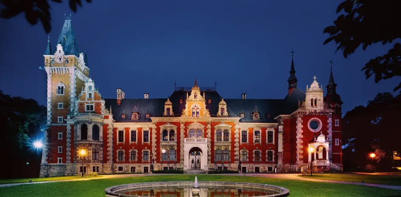 Pałac w Pławniowicach nocą, z oświetloną fasadą, wieżą zegarową i fontanną. Atrakcje tego miejsca zachwycają o każdej porze.
