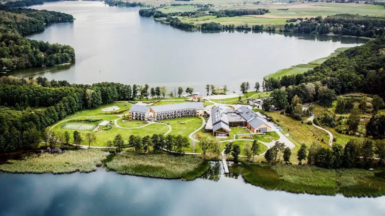 Malownicze mazury miejscowości nad jeziorem. Hotel z kortem tenisowym i plażą, otoczony zielenią i wodą.