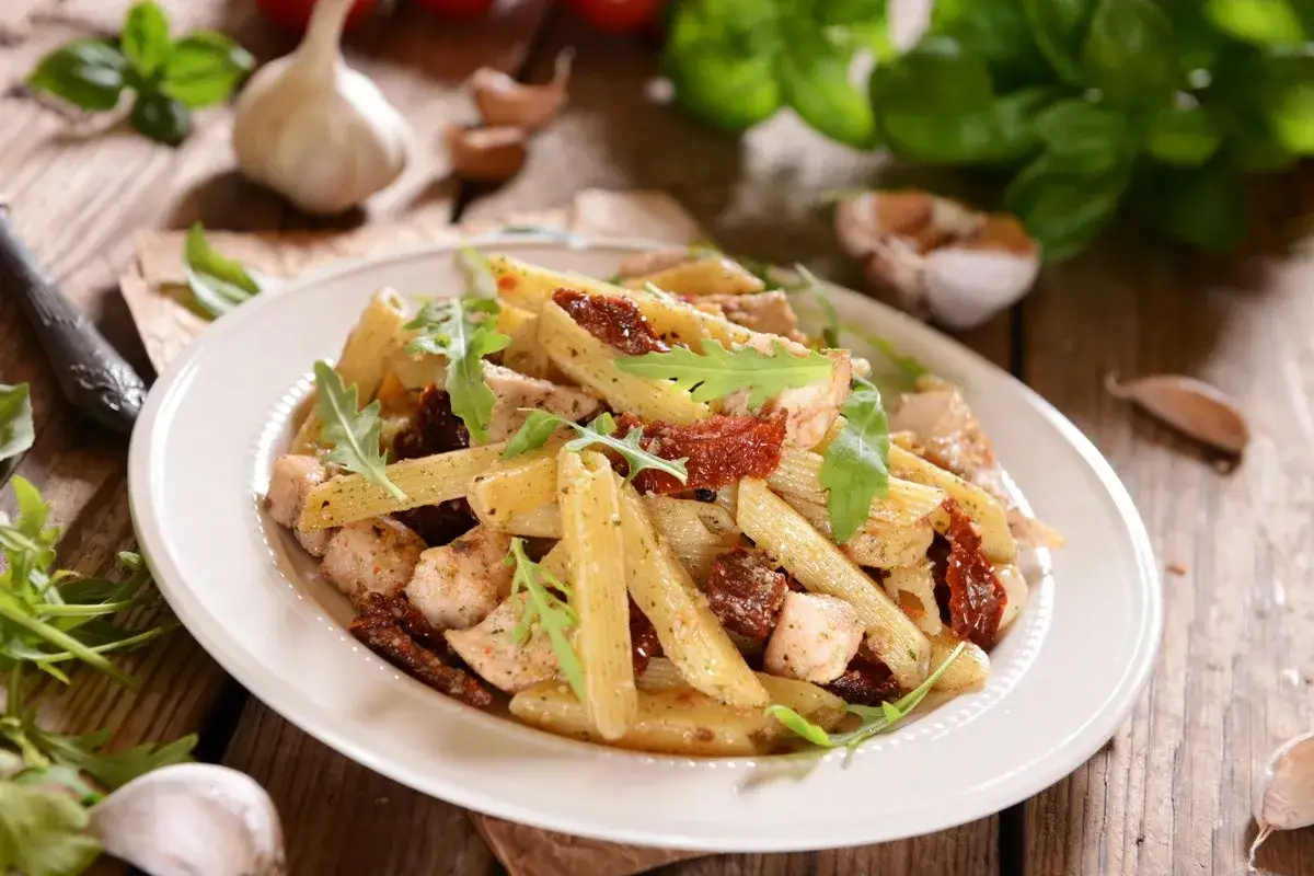 Penne z kurczakiem i suszonymi pomidorami, ozdobione świeżą rucolą. Idealne danie na szybki obiad.