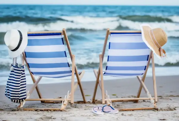 Dwa leżaki plażowe z niebiesko-białymi pasami, kapelusze i torba na piasku, z widokiem na morze.