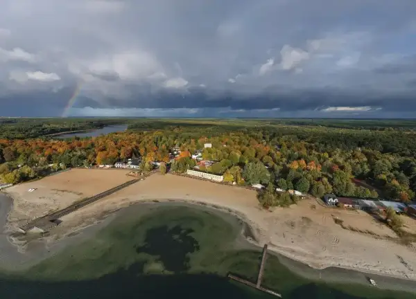 Jesioro Niedzięgiel w jesiennej odsłonie. Tęczowy łuk nad ciemnymi chmurami, piaszczysta plaża i kolorowe drzewa tworzą malowniczy krajobraz.