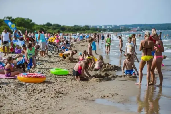 Tłum ludzi cieszy się słońcem i zabawą na plaży w Gdańsku. Dzieci budują zamki z piasku, a dorośli odpoczywają nad morzem.